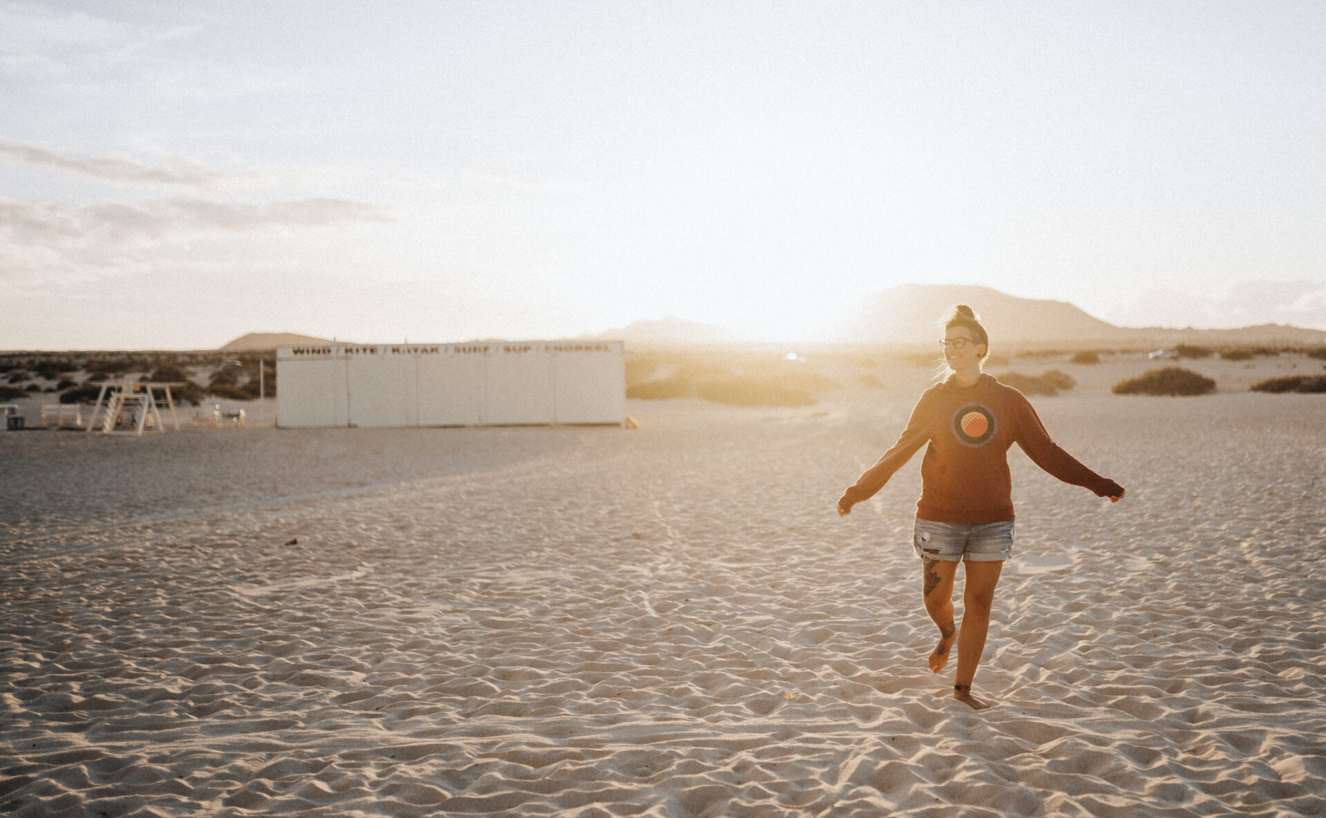 Gegenlichtaufnahmen beim Porträt Foto von Dominic Wolf, zu sehen ist Lisa Ludwig am Strand von Corallejo auf Fuerteventura während sie im Sand mit dem roten Pullover und der Jeansshorts tanzt