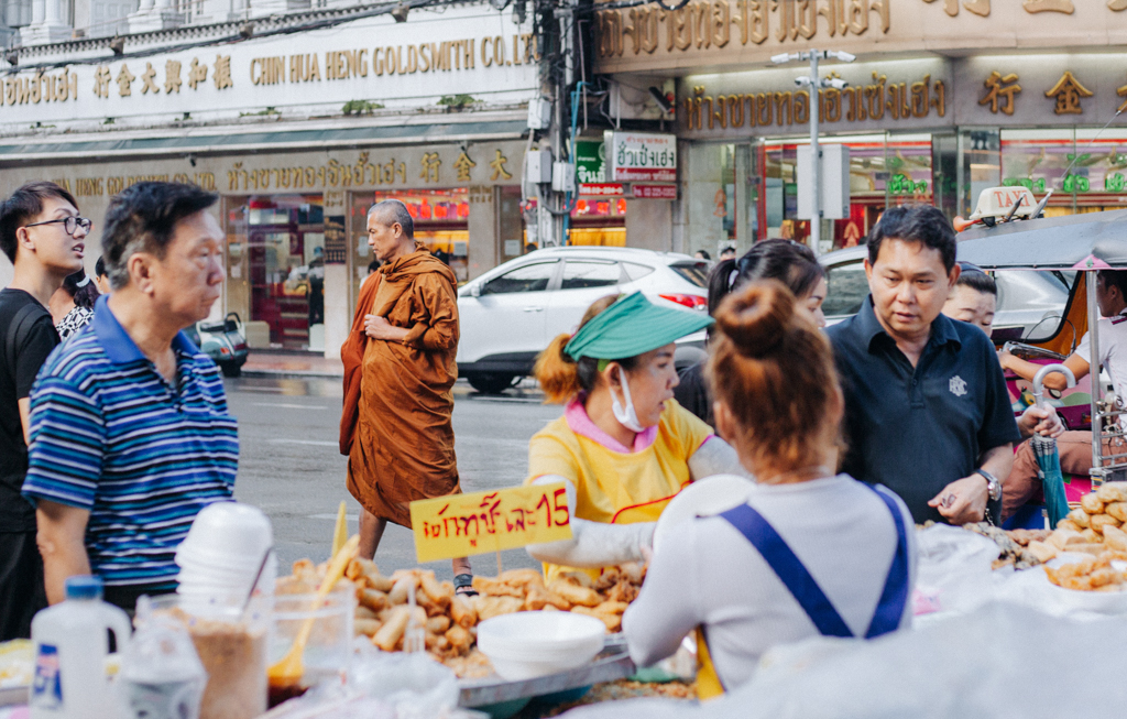 Aufnahme in China Town in Bangkok: ein buddistischer Mönch mit orangefarbigem Gewand läuft durch die Strasse während im Vordergrund Menschen an einem Essensstand etwas kaufen oder bestellen