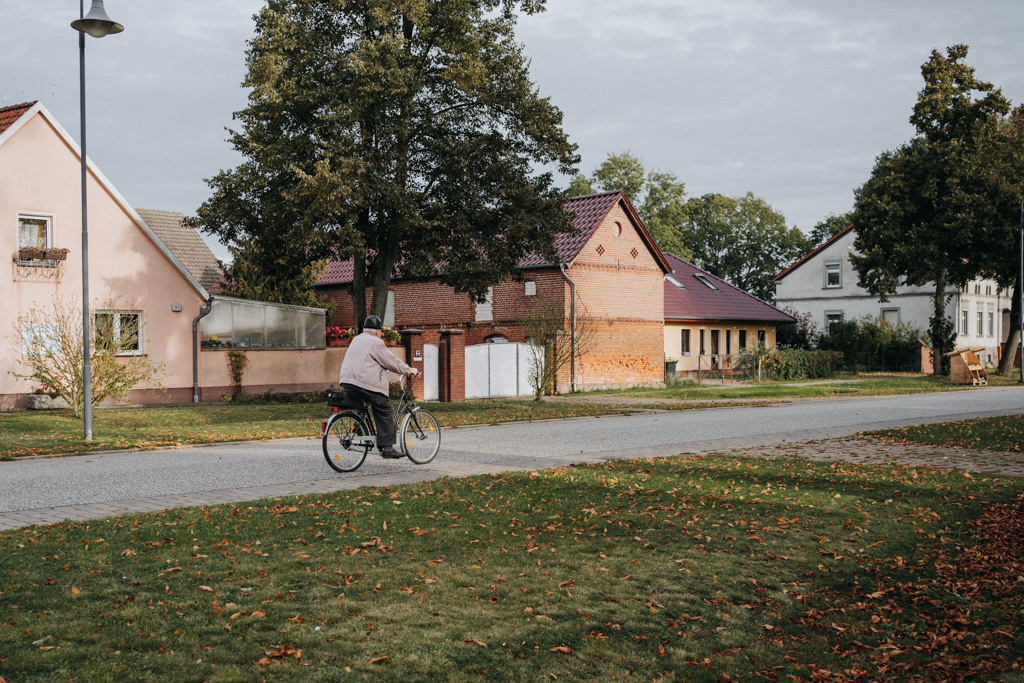 Streetfotografie: Ein alter Mann fährt mit seinem Fahrrad auf einer einspurigen Strasse in Brandenburg