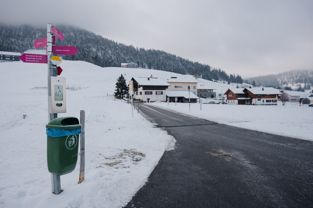 Ein Winterwanderweg mit Beschilderung, darunter Gassibeutel für den Kot von Hunden und ein grüner Mülleimer, Blick auf Hittisau mit schneebedeckter Wiese