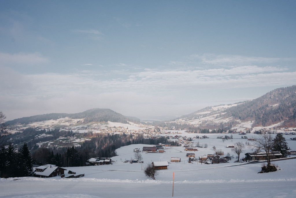 Blick beim Umgang in Hittisau auf das Dorf Hittisau im Bregenzerwald, schneebedeckte Wiesen und ein strahlend blauer Himmel