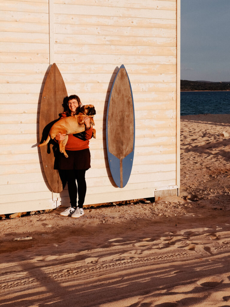 Lisa hält Frau Maier hoch am Strand vor Surfbrettern, Sardinien mit Hund