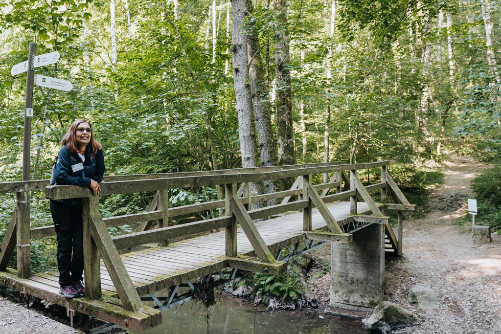 Wandern in der Vulkaneifel immer an der kleinen Kyll entlang, Deepti erklärt die nächste Übung mit dem Wasser